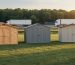 Three backyard sheds made of wood, metal, and resin aligned diagonally on a grassy lot at golden hour, with a tree line, a distant road with a semi-truck, and muted cargo containers in the background.