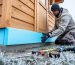 Worker fitting blue rigid foam boards to the exterior foundation of a wooden shed with excavated soil and frosty grass, under cool overcast daylight.