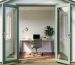 Eye-level view into a converted backyard shed home office with open double doors, a desk and ergonomic chair against a subtle geometric wallpaper accent wall, plants and task lamp inside, and a softly blurred garden and fence beyond.