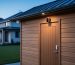 Backyard shed with an outdoor security camera above the door and a wireless door sensor, shot at dusk from a 45-degree angle, with the main house and discreet outdoor Wi‑Fi node blurred in the background.