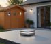 Smart home hub with glowing indicator on an outdoor table, with a wooden garden shed and house in the background; small door sensor visible on the shed door under warm evening light, shallow focus.