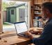 Small business owner typing on a laptop at a neat workshop bench, pegboard tools and storage bins visible, daylight illuminating the space, and a modern backyard storage shed seen through an open doorway; laptop screen out of focus.