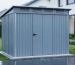 Modern metal weatherproof storage shed in a rainy backyard with water beading on panels, elevated foundation and roof vents visible, wet lawn and garden beds softly blurred in the background.