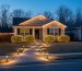 Suburban house at twilight with warm white LED lights along the roofline and fence, net lights on shrubs, and solar-powered pathway stakes illuminating a stone walkway; neighboring homes and bare trees softly blurred in the background.