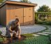Eco-friendly modern backyard shed on compacted gravel near a property edge with a homeowner kneeling to test soil using a screwdriver, mature trees and native plantings softly blurred in the background at golden hour.