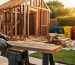 Builder aligning a reclaimed board on a partially built shed frame, with blurred background showing sorted recycling bins, stacked salvaged wood, and garden in warm golden hour light.