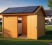 Eye-level wide shot of a wooden backyard shed with solar panels on the roof and a wall-mounted battery visible through a half-open door, lit by warm evening sunlight, with lawn, garden beds, trees, and a distant house softly blurred in the background.