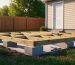 Interlocking concrete deck blocks arranged under pressure-treated beams forming a level shed foundation on a slightly sloped backyard, photographed at golden hour with a softly blurred fence, trees, and house in the background.