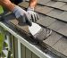 Gloved hands of a homeowner on a ladder spreading roofing cement with a putty knife on a leaking asphalt-shingle shed roof next to the gutter, with a blurred backyard and tools behind.