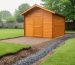 Backyard wooden shed on a graded gravel pad with a shallow gravel swale channeling water away, under bright overcast light with lawn, fence, and trees in the background