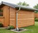 Eye-level three-quarter view of a wooden backyard shed featuring K-style aluminum gutters and a downspout draining onto a stone splash block, with raindrops on the metal and a wet lawn and fence softly blurred in the background.