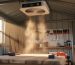 Ceiling-mounted air filtration unit operating in a wooden shed workshop, with dust particles visible in sunlight and a workbench and hand tools in the background.