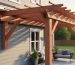 House-attached cedar-tone wooden pergola anchored to a ledger board on gray fiber-cement siding, shading a small patio in warm evening light, with patio chairs, planters, and lawn softly blurred in the background.