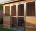 Backyard wooden shed with stainless steel mesh security screen door and matching window screen, golden-hour side light highlighting the sturdy frame; blurred garden and fence behind, with a lawn mower faintly visible inside.