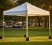 White 10x10 pop-up canopy with aluminum frame and leg weights on a grassy park lawn at golden hour, with blurred trees and market tables behind it.