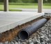 Low-angle view of a carport slab edge with an exposed French drain trench, showing perforated pipe wrapped in fabric and surrounded by gravel, with water draining away and the carport and driveway softly blurred in the background.