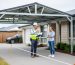 Building inspector with clipboard discusses a partially installed metal carport with a homeowner beside a suburban house; survey stakes and a string line mark setbacks, with driveway, shrubs, and a neighboring fence in soft background.
