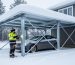 Building inspector in winter gear examines a cross-braced steel carport bearing thick snow, with a car parked underneath and a snowy suburban home and pines in the background under overcast light.
