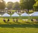 Row of pop-up canopy tents in different sizes, from small to large, set side-by-side on a grassy park field at golden hour with proportional seating and a few people under each; trees and a walking path softly blurred in the background.