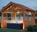 Evening view of a cedar gazebo with a vented gable roof, open louvers, and a ceiling fan above a steaming hot tub, warm lights inside and a landscaped backyard softly visible behind.