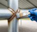 Gloved hand brushing gray zinc-rich primer onto a rusted welded joint of a galvanized portable garage steel frame, with the shelter’s fabric cover and gravel driveway softly blurred behind.