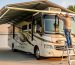 Person on a step ladder measuring the height of a steel RV carport as a Class A motorhome approaches, showing generous overhead clearance at golden hour