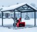 Sturdy snowblower shelter with a pitched, snow-covered roof on a reinforced concrete pad, steel frame and anchor bolts visible, red snowblower parked inside, with a blurred suburban house and snowy trees in the background.