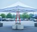 White canopy tent on asphalt secured with heavy leg weights and ratchet straps to concrete blocks, with a market parking lot and trees softly blurred in the background.