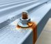 Close-up of a steel screw on an aluminum carport roof showing rust streaks, white powdery corrosion, and water droplets, with blurred carport beams and driveway in the background.