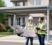 Homeowner and building inspector in a hard hat review rolled plans next to a modern metal carport in a suburban driveway, with the house and trees softly blurred in the background under diffused morning light.