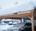 Shallow-pitched suburban carport roof burdened by deep snow with slight beam sagging, a parked car beneath, and a snow-covered driveway and trees in the background on an overcast day.