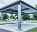 Low-angle three-quarter view of a modern metal carport featuring heavy-gauge galvanized, powder-coated steel frame and base plates anchored to concrete footings, with ribbed roof panels and a suburban driveway background.