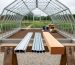 Aluminum extrusion, galvanized steel tube, and cedar beam on a wooden sawhorse in front of a partially assembled greenhouse frame, with blurred gravel bed and concrete footing in a garden setting under soft daylight.