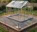 Aluminum-frame hobby greenhouse under construction on compacted gravel with timber edging, corner concrete piers and anchor brackets, stakes and level string lines in a backyard at golden hour.