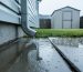 Ground-level view of rainwater pooling against a home’s concrete foundation beside a downspout, a faint crack along the nearby patio slab, and a slightly leaning backyard shed blurred in the background under overcast light.