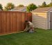 Homeowner kneeling in a suburban backyard measuring from a wooden privacy fence to a small garden shed with a yellow tape measure at golden hour, with neighboring house, lawn, and trees in the background.