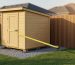 Portable wooden shed on skids over a gravel pad with a yellow tape measure extending from the shed corner to a wooden privacy fence, in warm late-afternoon light with a suburban yard and house roofline softly blurred in the background.