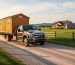Flatbed truck transporting a wooden storage shed on a rural gravel road toward a farmhouse, with a mailbox, pasture, fence line, and distant hills in warm golden hour light.