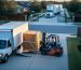 Forklift unloading a crated wooden shed from a small box truck onto a narrow suburban driveway, with a long semi-truck waiting near a gated entrance, illustrating limited access delivery.