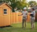 Building inspector measuring setback distance to a wooden shed on skids in a suburban backyard while the homeowner observes, with fence, trees, and neighboring houses softly blurred in the background