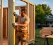 Carpenter measuring a 2x4 stud on a partially framed wooden shed at golden hour, with sharp structural details and a suburban backyard softly blurred in the background.