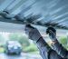 Low-angle view inside a metal carport showing a worker attaching a white anti-condensation membrane to the underside of corrugated roof panels, with water droplets on uncovered metal and a car parked below in soft focus.