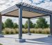 Aluminum pergola with louvered roof and retracted canopy securely anchored to concrete footings on a paver patio, with wind-swept grasses, trees, and fast-moving clouds in a suburban backyard.