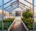 View from the greenhouse doorway showing polycarbonate panels, roof vents open, bubble-wrap insulation on the north wall, and zoned plantings—tomatoes and peppers in the sunny south beds, greens at center, and lettuce/spinach along the cooler north row—with soft morning light and a lightly frosted yard outside.