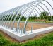 Steel quonset greenhouse hoops connected to an anchored concrete foundation with visible base plates and anchor bolts, showing drainage-friendly site grading.