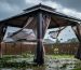 Wind and storm rain whip torn fabric on a damaged backyard gazebo, with the canopy lifted and distorted by strong gusts.