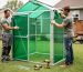 Two people work on assembling a 6x8 greenhouse frame in a backyard, with a wall panel held in place and tools nearby.