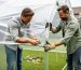“Two people assembling a portable greenhouse frame and stretching a clear plastic cover in a backyard”