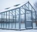 Snow-covered greenhouse with a metal frame in a winter backyard under overcast daylight.