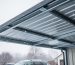 Metal carport roof under light snowfall, viewed from below to show roof beams and snow coverage.